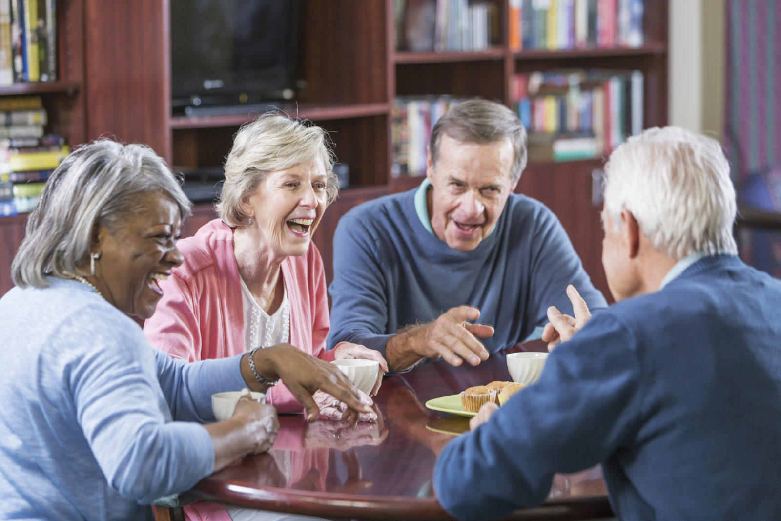 A multi-ethnic group of senior men and women sitting at a table, conversing while having a snack and tea or coffee.