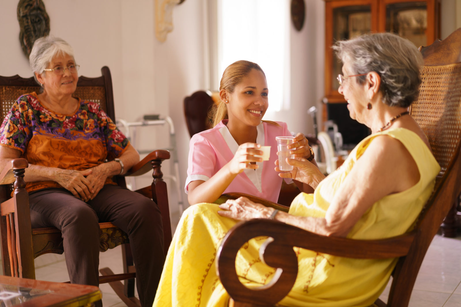 Old people in geriatric hospice: young attractive hispanic woman working as nurse helps a senior woman. She gives a water glass and prescription medicine to the aged patient