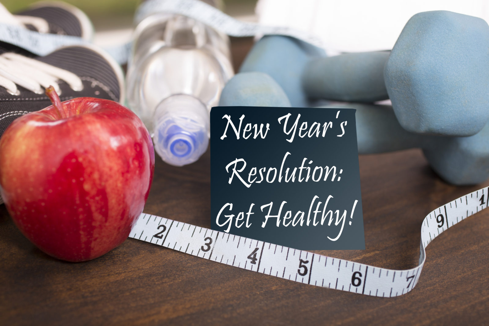 New Year's Resolution to get healthy in January of the coming year. Image features: dumbbells, sports shoes, water bottle, apple, towel, tape measure on wooden table. Adhesive note reading "New Year's Resolution: Get Healthy!" in foreground.